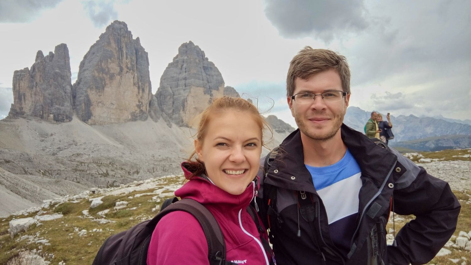 A woman in a pink jacket and a man in a black jacket smile while taking a selfie in front of the Tre Cime di Lavaredo, three prominent mountain peaks in the Dolomites. The backdrop features rocky terrain and cloudy skies. Other hikers can be seen in the distance.