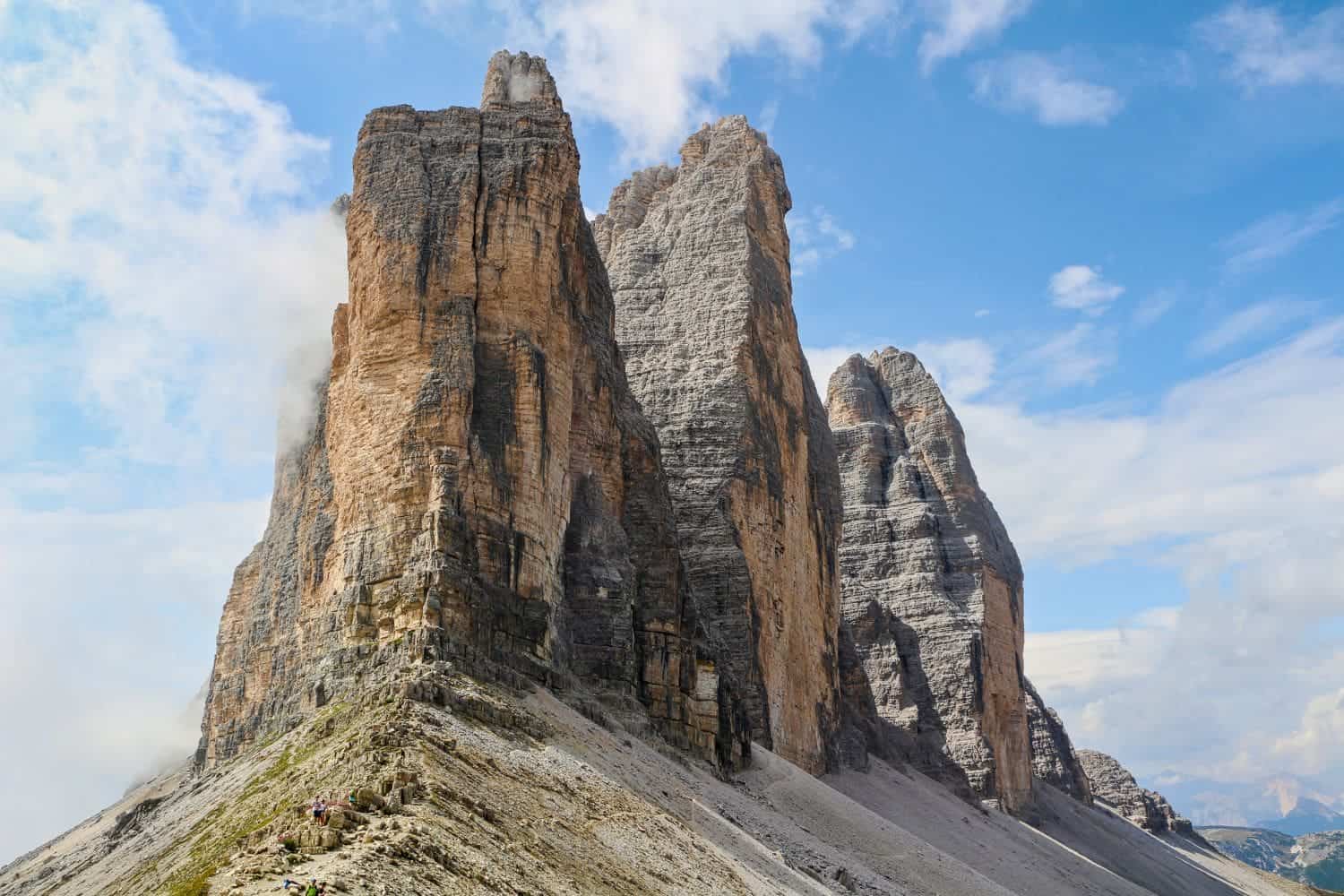 A rugged mountain landscape featuring three towering peaks known as Tre Cime di Lavaredo, set against a clear blue sky with a few clouds. The rock formations are steep and jagged, and the surrounding area is rocky with some green patches.