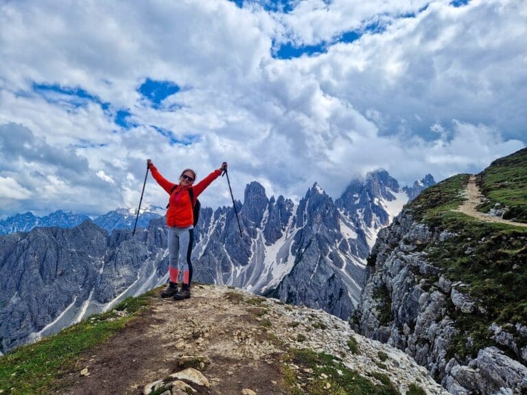 A person in a red jacket stands triumphantly on a rocky mountain edge, holding hiking poles aloft. Snow-capped peaks and a partly cloudy sky form a dramatic backdrop.