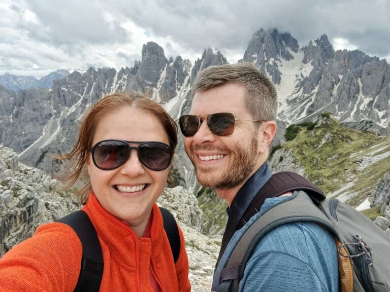 A smiling woman and man take a selfie on a mountainous hiking trail with jagged peaks and patches of snow in the background. They are wearing backpacks and sunglasses. The sky is cloudy.