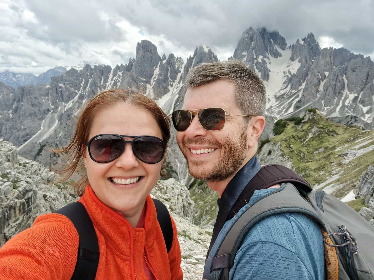 Are the Dolomites worth visiting? Yes! Why? 9 A smiling woman and man take a selfie on a mountainous hiking trail with jagged peaks and patches of snow in the background. They are wearing backpacks and sunglasses. The sky is cloudy.