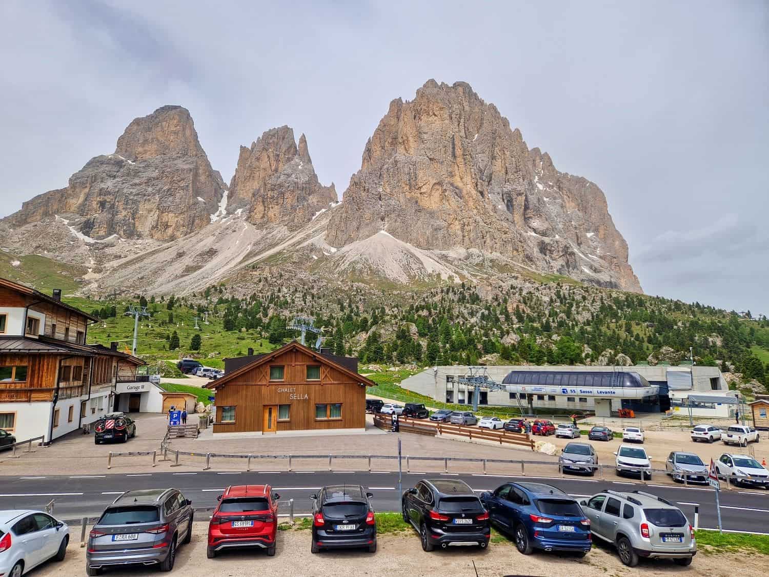 Are the Dolomites worth visiting? Yes! Why? 6 A scenic view of towering rocky mountains under a cloudy sky. In the foreground, a row of parked cars lines a road in front of wooden buildings. Green trees dot the landscape, adding contrast to the rugged mountain backdrop.