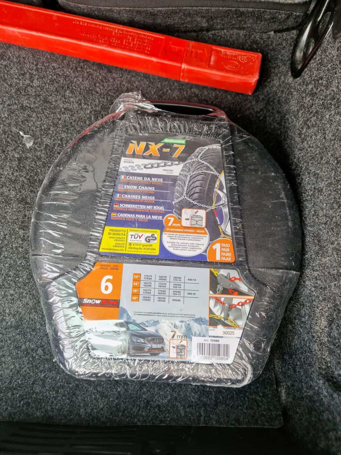 A pack of NX-7 snow chains for tires, still wrapped in plastic, lies in a car trunk. The packaging indicates compatibility with 6 tire sizes and features illustrations of a tire and snow. A red object is visible nearby.
