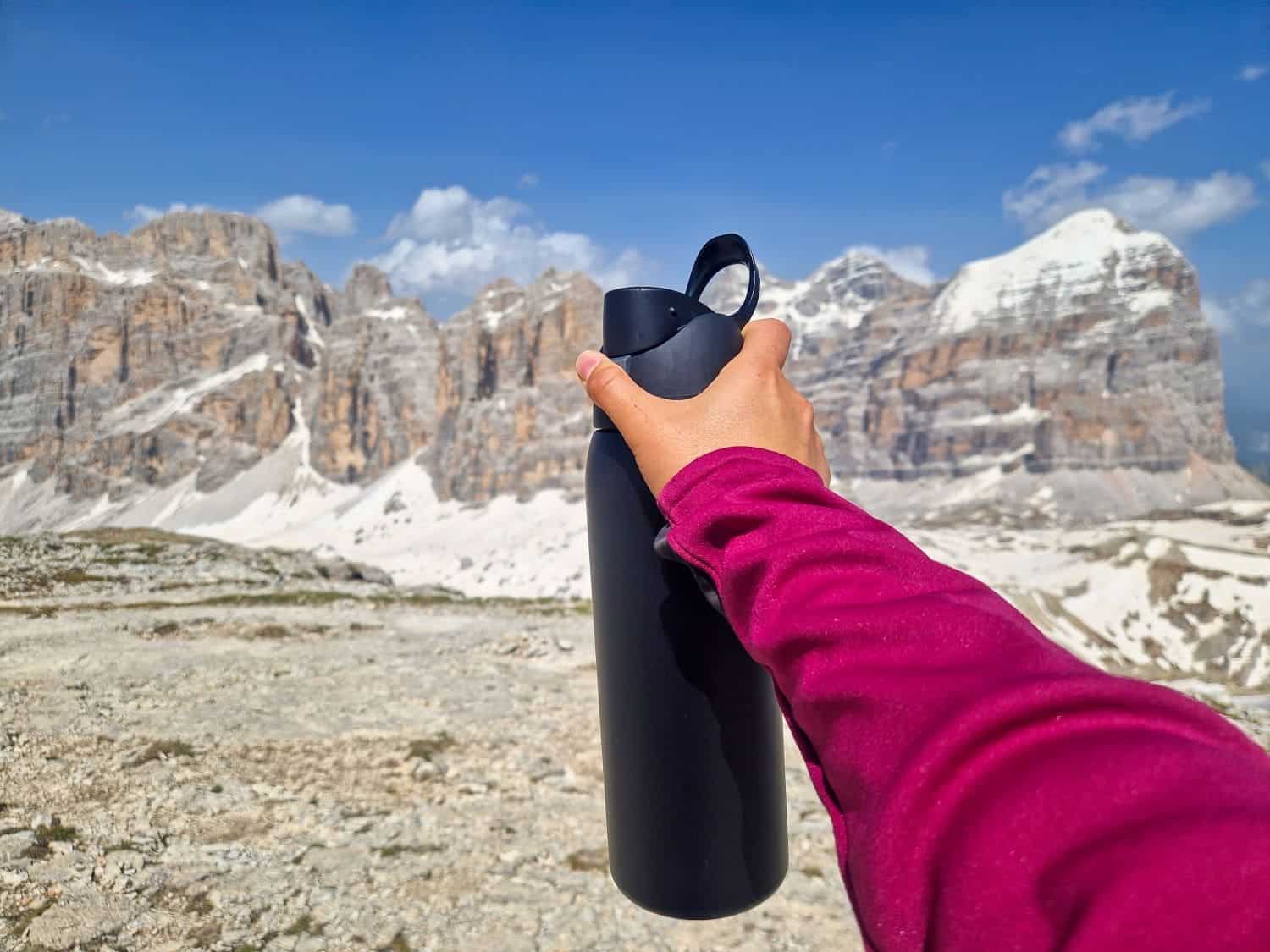 A person wearing a pink jacket holds a black water bottle in front of a scenic mountain range with snowy peaks under a clear blue sky.