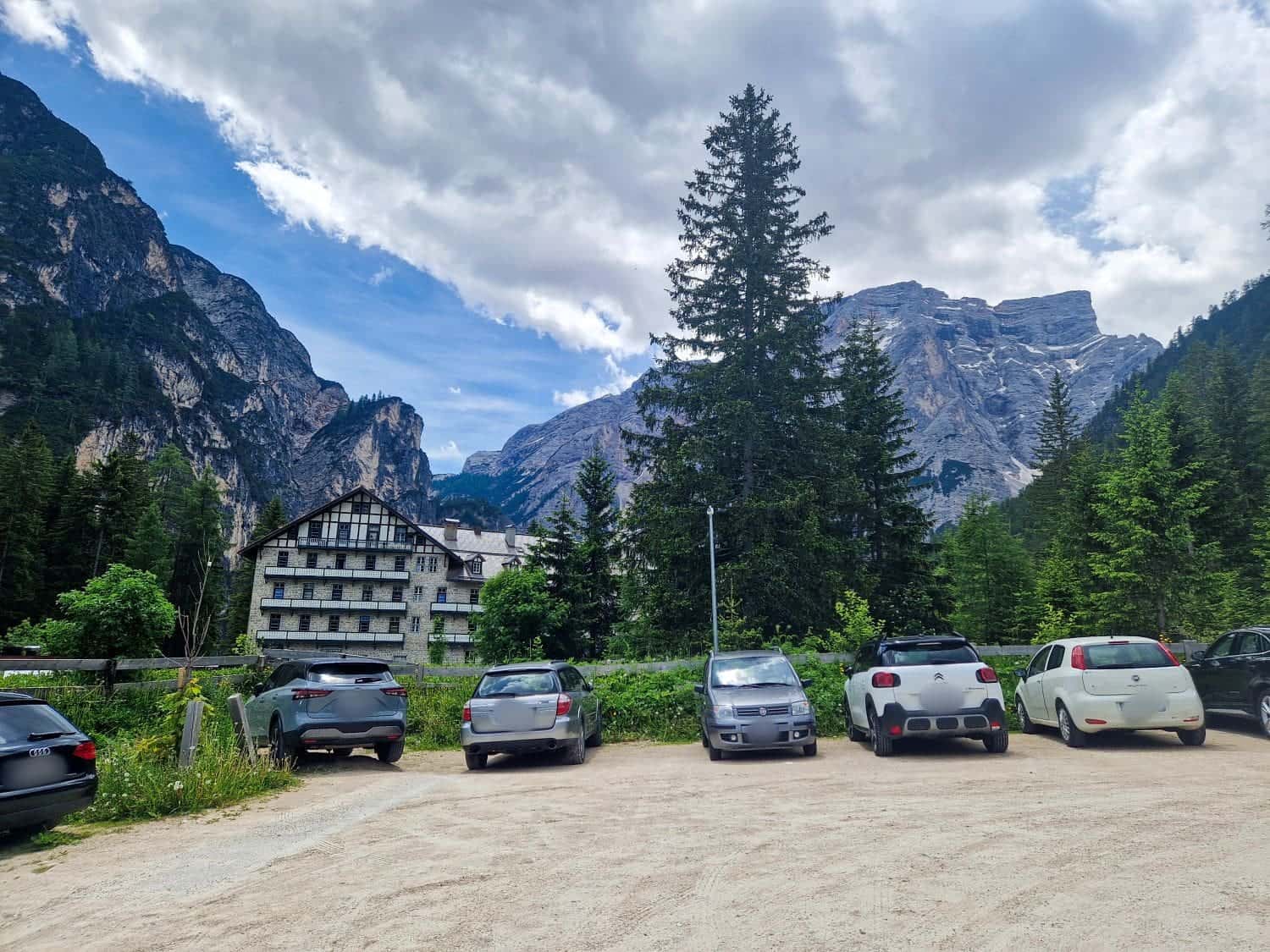Lago di Braies: Discover The Pearl of the Dolomites 5 A parking lot with several cars is in the foreground. Behind, there are tall evergreen trees and a rustic building. In the background, majestic mountains rise under a cloudy sky, creating a picturesque natural setting.