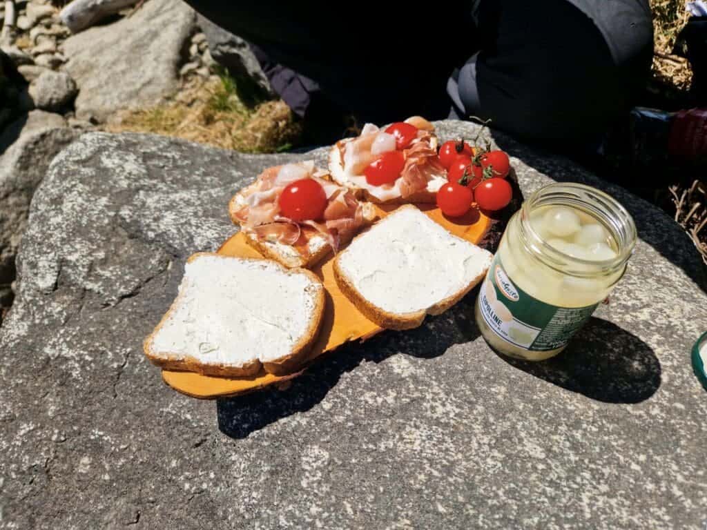 A picnic setup on a rocky surface featuring slices of bread with spread, prosciutto, cherry tomatoes, and a jar of pickled onions. A person sits nearby, partially visible, enjoying the outdoor meal.