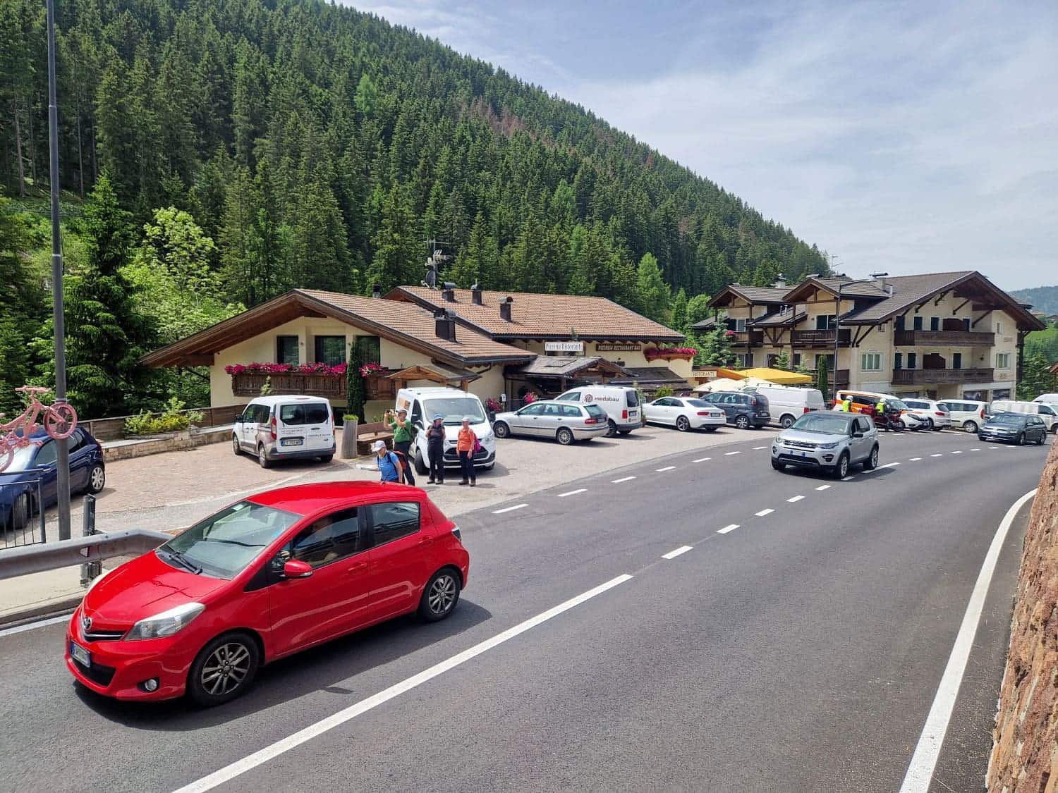 A scenic mountain road with a row of cars and vans parked near a rustic building. Dense green forest covers the hillside in the background, and a few people stand near the vehicles under a partly cloudy sky.