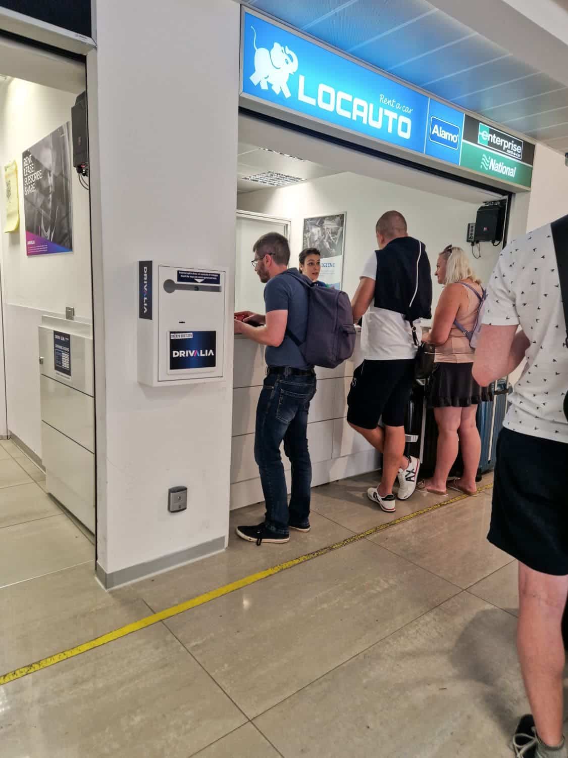 People are standing at a car rental counter labeled "Locauto," engaging with staff. A man with a backpack and a woman in a white shirt are in focus. The area is marked by a yellow line on the floor.