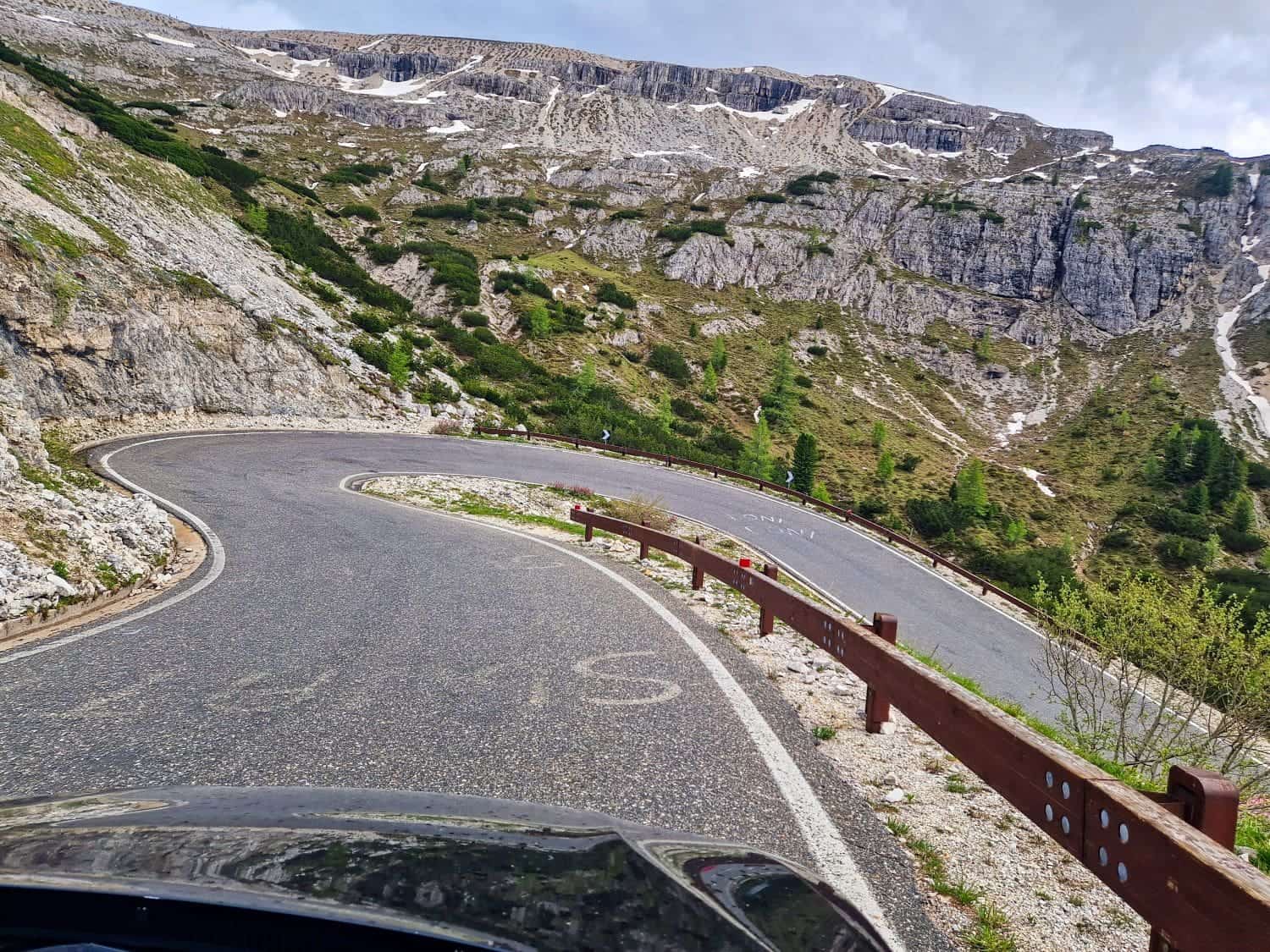 A winding mountain road curves sharply with rugged rock formations and green vegetation lining the route. The overcast sky adds a moody ambiance to the scenic landscape. A wooden guardrail follows the road's edge.