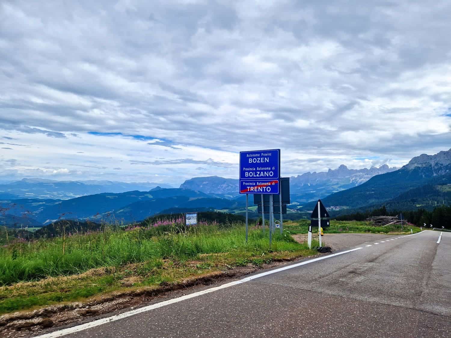 Driving in Dolomites road with a scenic view of a mountainous landscape with a road sign indicating directions to Bozen/Bolzano, Trento, and Brenner. The sky is cloudy, and the road curves through lush green fields and distant peaks.