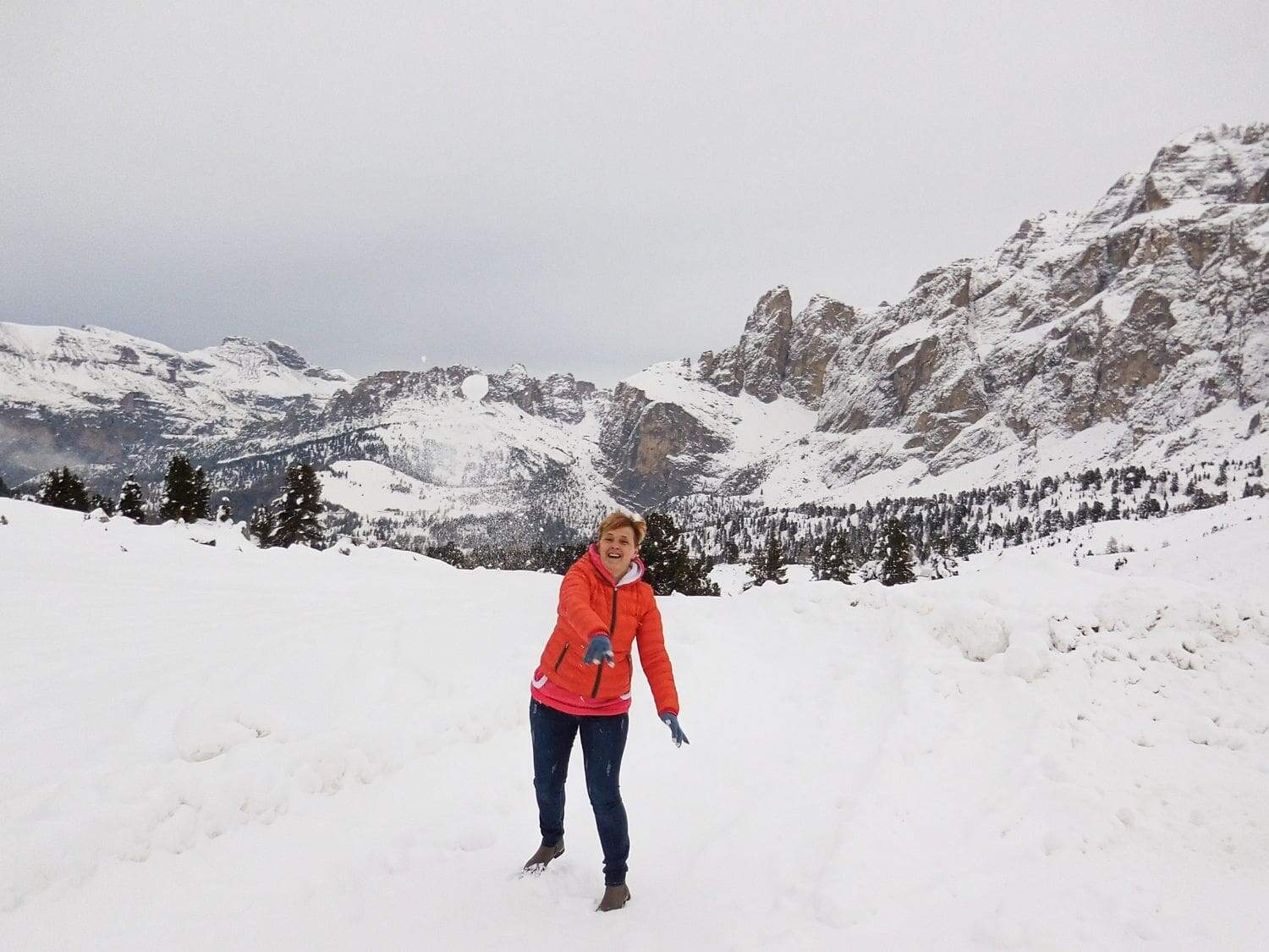 A person in a bright orange jacket stands joyfully in a snowy landscape, surrounded by tall mountains and trees. The sky is overcast, enhancing the wintery atmosphere.