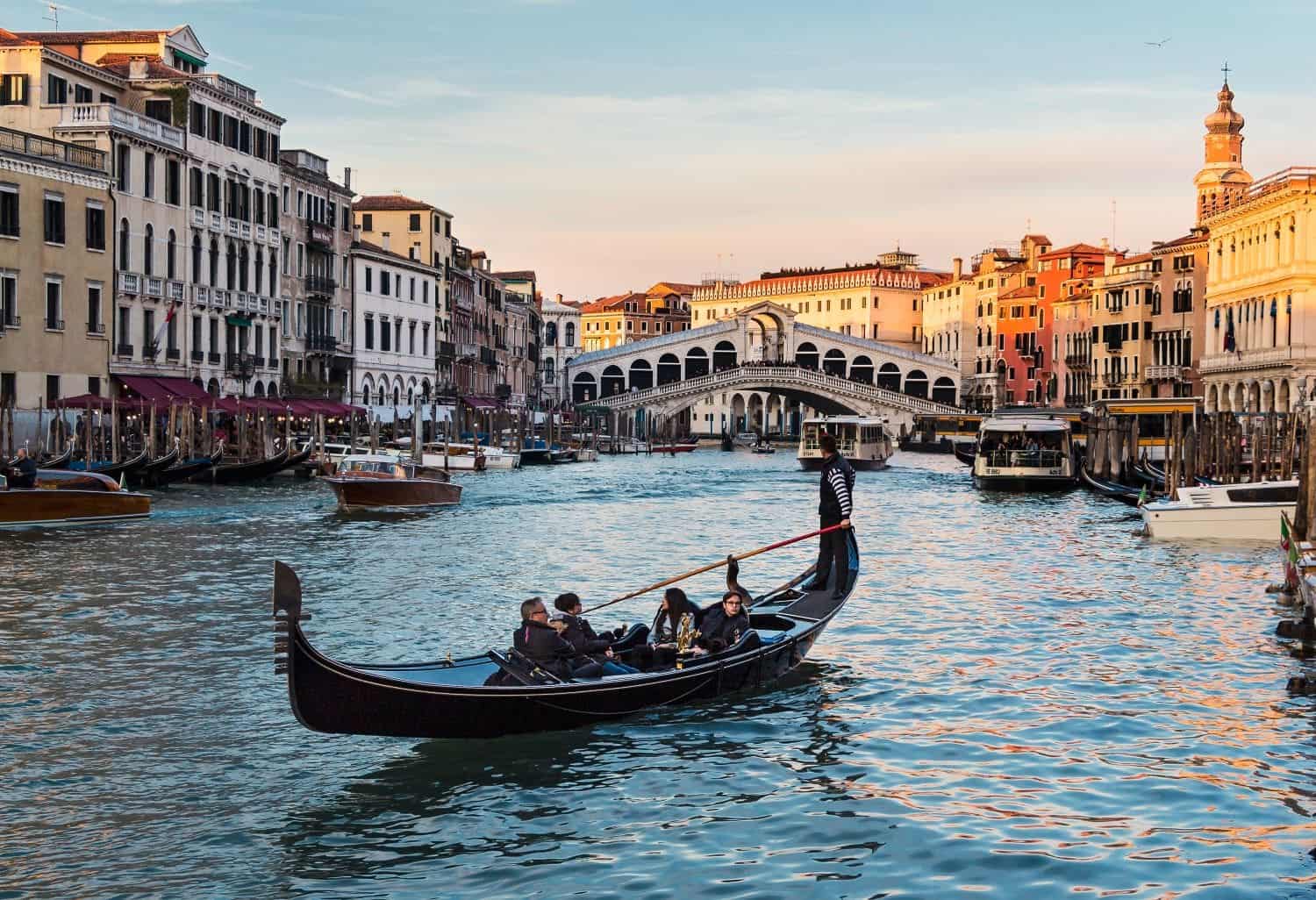 A gondola with passengers glides along a canal in Venice, surrounded by colorful buildings. The Rialto Bridge is visible in the background, and the sun casts a warm glow over the scene.