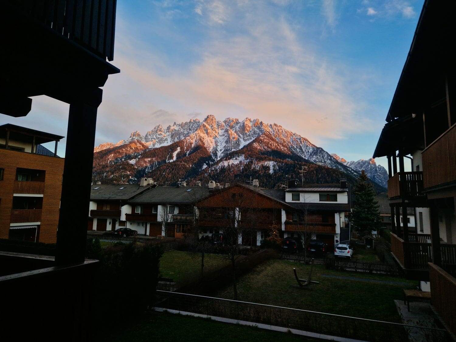 Are the Dolomites worth visiting? Yes! Why? 14 A scenic view of snowy mountains during sunset, with orange and pink hues reflecting on the peaks. In the foreground, several houses and buildings are silhouetted against the vibrant sky.