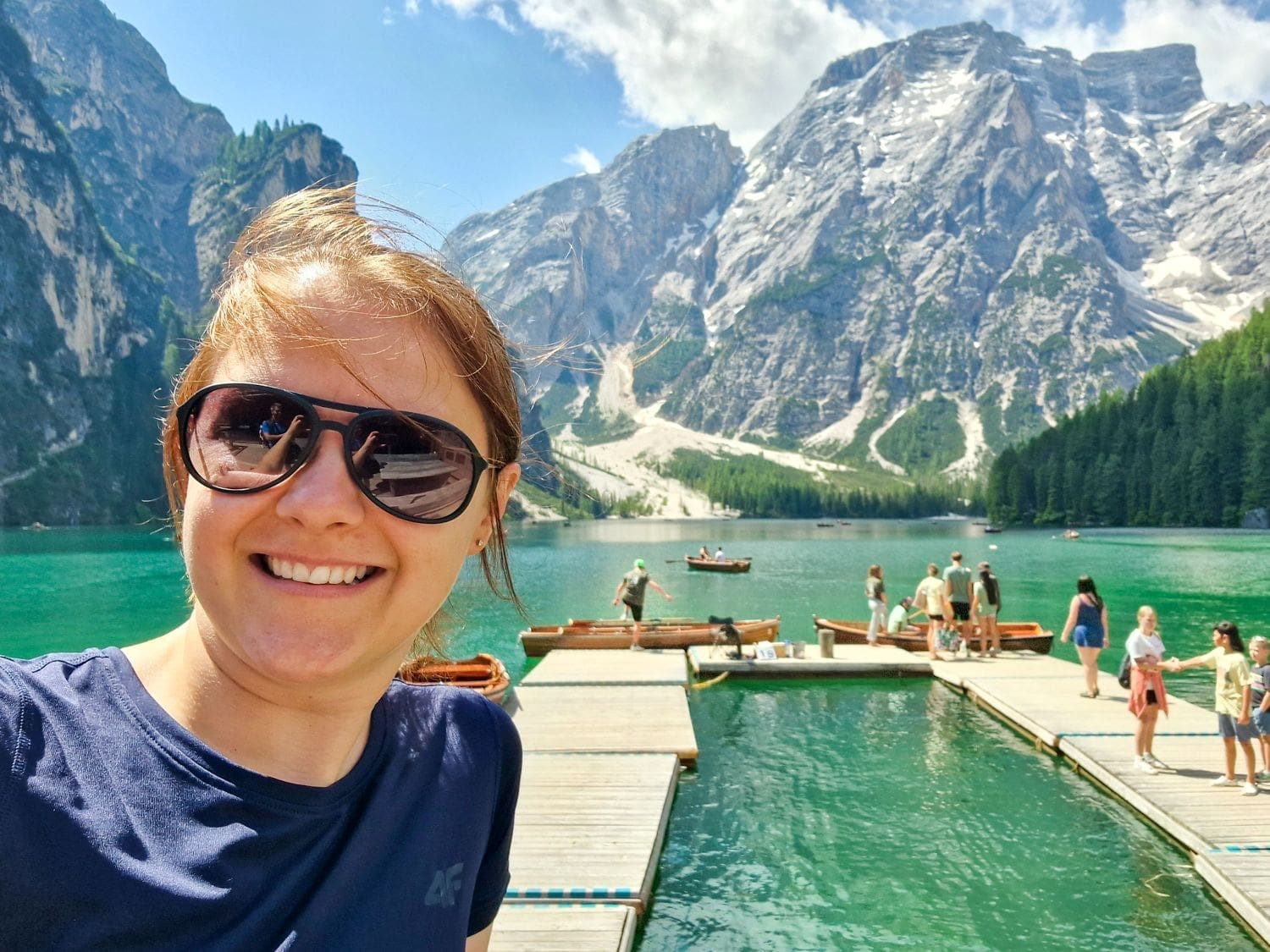 A person wearing sunglasses is taking a selfie in front of a scenic mountain lake Lago di Braies, one of the reasons the Dolomites are worth visiting. Behind them, people are standing on wooden docks, and there are boats on the water. The backdrop features tall mountains and a partly cloudy sky.