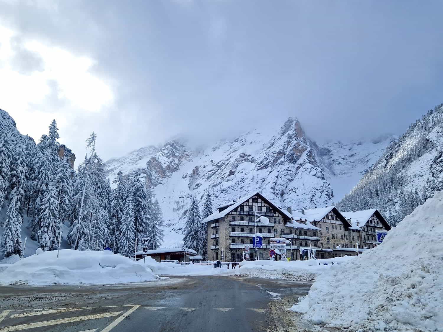 Lago di Braies: Discover The Pearl of the Dolomites 16 A snowy mountain scene with snow-covered buildings and trees. The sky is overcast, and mist partially obscures the mountain peaks in the background. The road in the foreground is clear with visible snow piled on the sides.