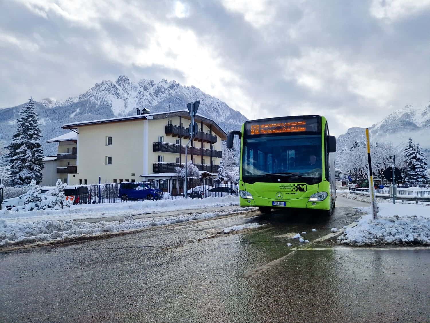 A green bus travels on a snowy road in a mountainous area. Snow-covered buildings and trees line the street, with dramatic snow-capped peaks visible in the background under a cloudy sky.