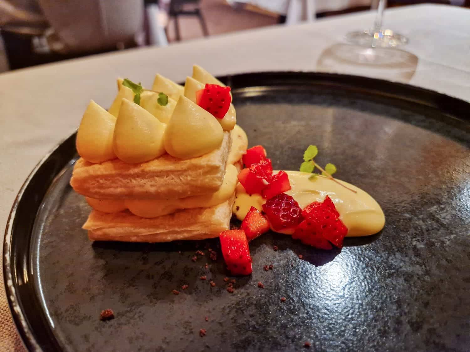 A dessert of layered puff pastry topped with cream dollops, garnished with red berry pieces and a mint leaf, served on a glossy black plate.