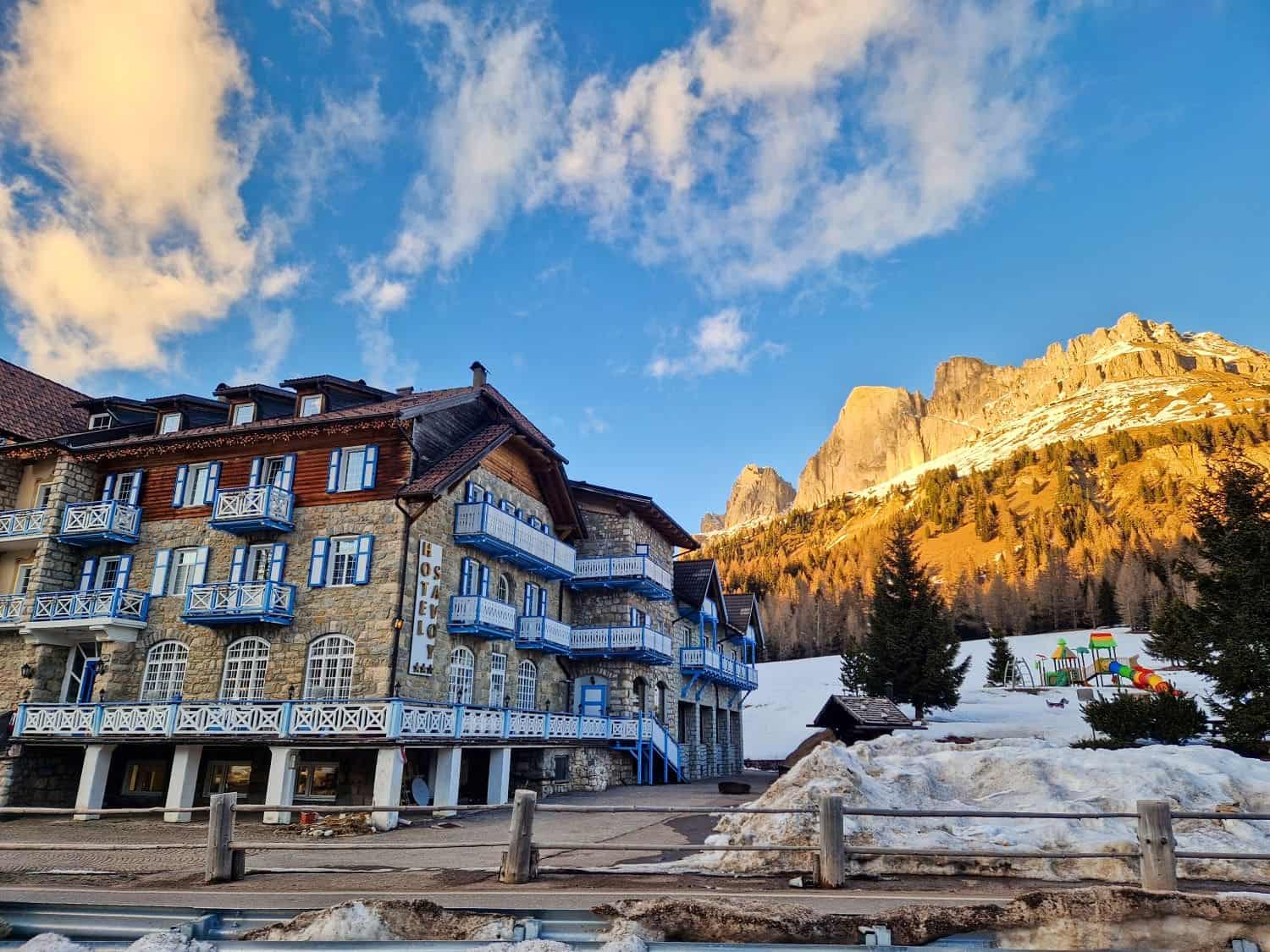 A charming stone hotel with blue balconies sits beside snow-covered ground, overlooking a rugged mountain bathed in golden sunlight. The sky is clear with scattered clouds, and a playground is visible in the background.