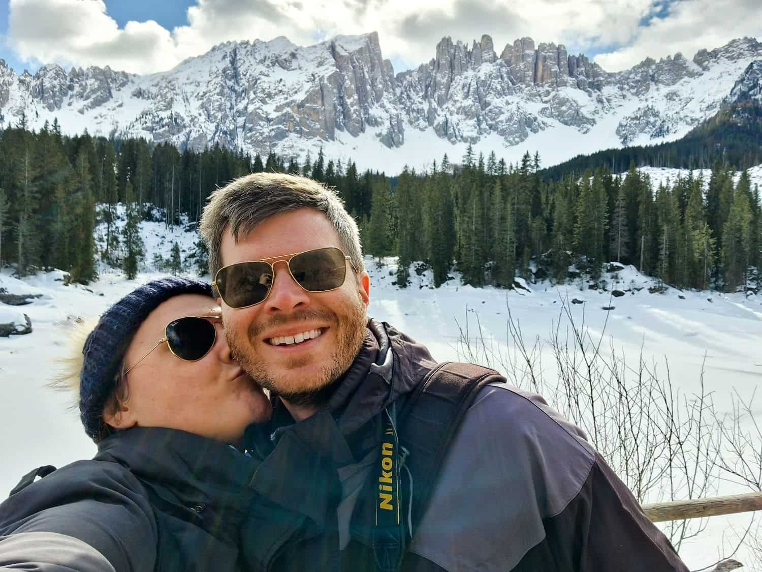 Are the Dolomites worth visiting? Yes! Why? 16 A couple dressed in winter jackets pose for a selfie in a snowy landscape. The woman kisses the man's cheek. Behind them are snow-covered trees and a mountain range under a partly cloudy sky. The man has a camera strap around his neck.