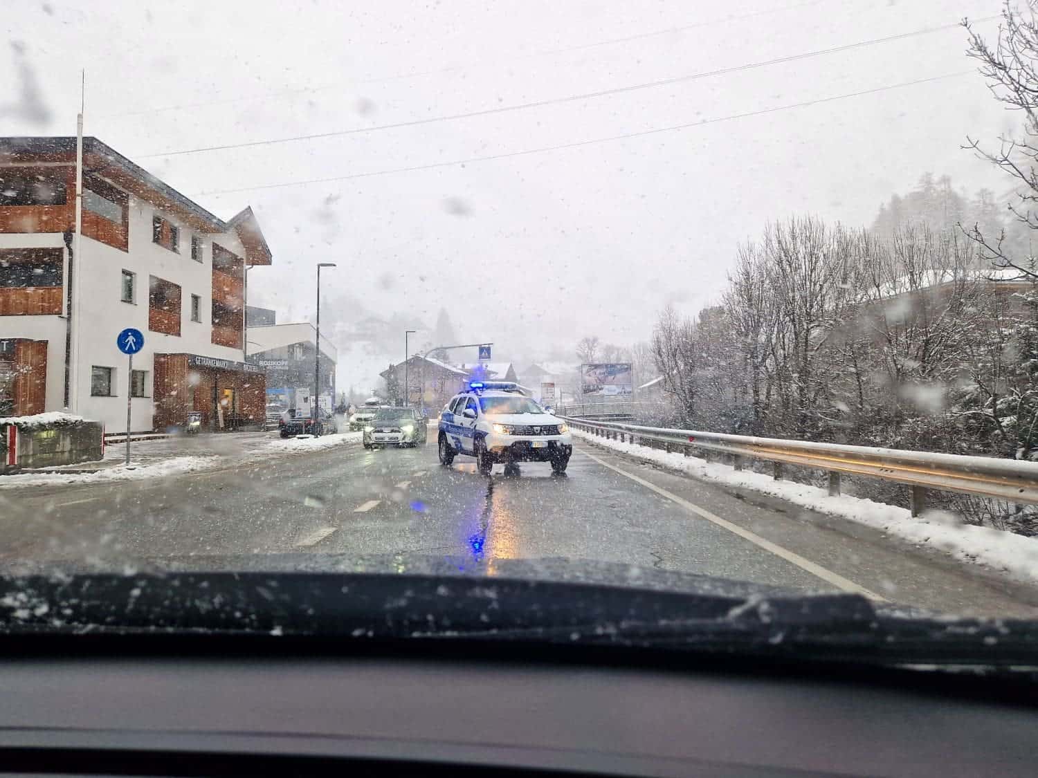 A snowy road seen from inside a car, with a police vehicle blocking one lane. The scene shows buildings on the left, trees on the right, and a wintry atmosphere with snow on the ground and falling.