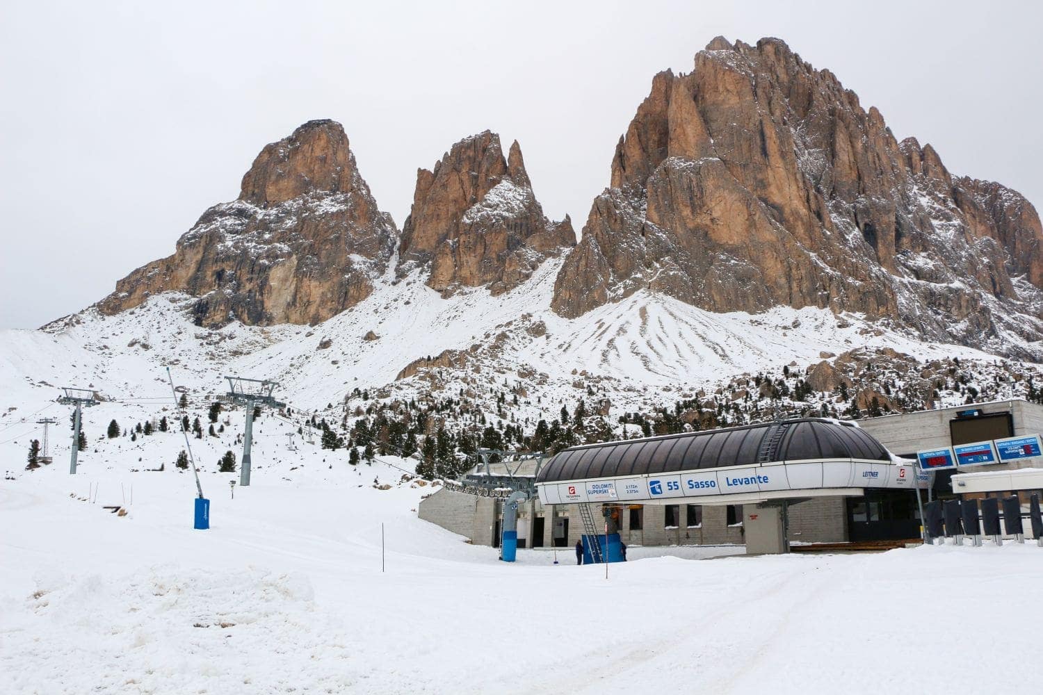 Snow-covered mountains with jagged peaks rise behind a ski lift station. The building has signs and blue accents, and ski trails can be seen leading away from it. The sky is overcast, creating a tranquil winter scene.