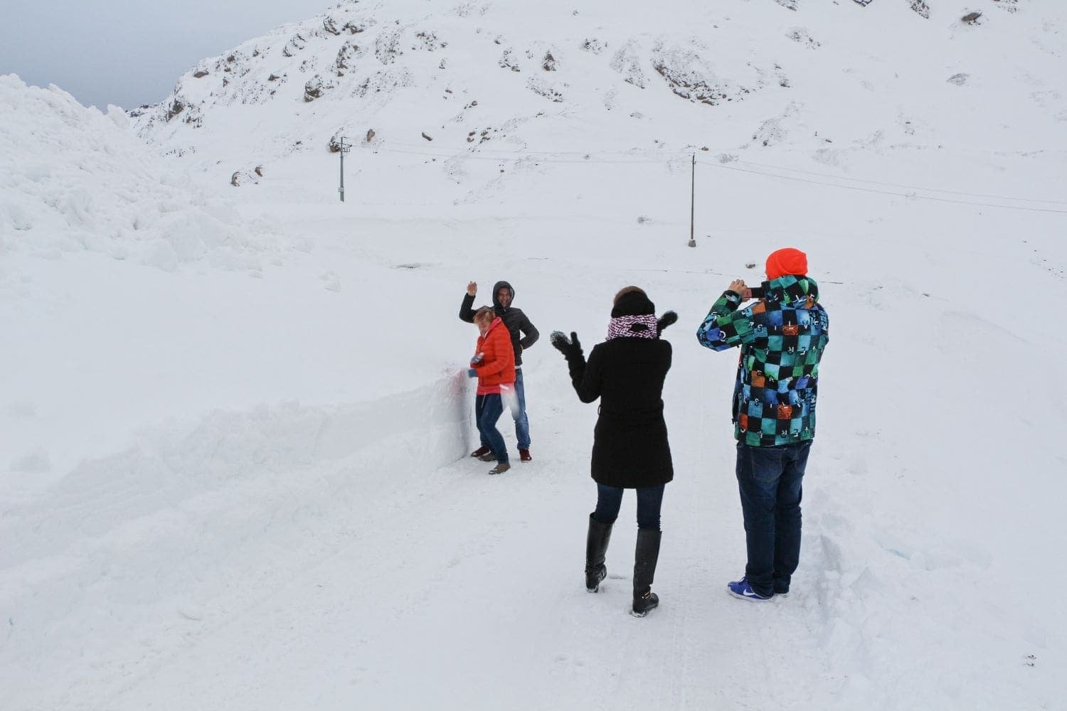 Four people stand on a snowy path surrounded by high snowbanks, dressed in winter clothing. Two face the camera, one raises a hand, and two face away, capturing the snowy landscape. Mountains and cloudy skies are in the background.