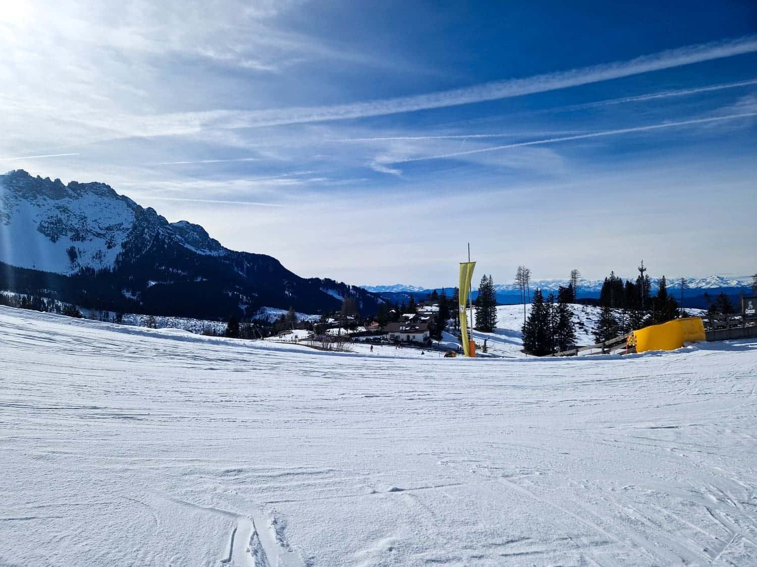 A snowy ski slope under a clear blue sky with mountains in the background. A small cluster of buildings and trees can be seen in the distance. Yellow barriers are scattered on the snow.