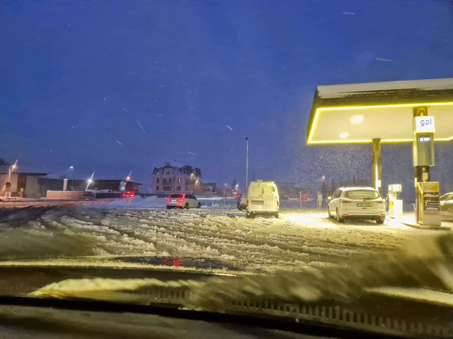 A snowy gas station scene at dusk, with several cars parked under illuminated fuel pump canopies. The ground is covered in snow, and lights from a nearby building and distant vehicles add a warm glow to the cold evening atmosphere.