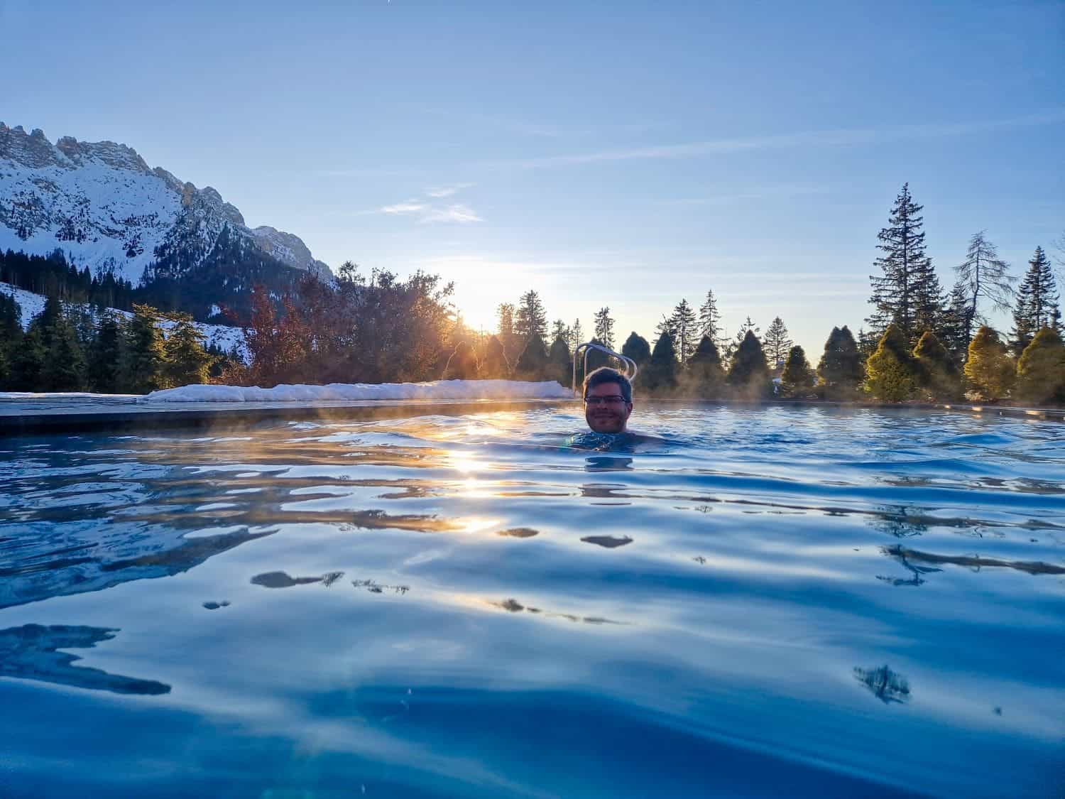 A person swimming in an outdoor pool with scenic mountains in the background. The sun is setting, casting a warm glow over the water. Snow is visible on the distant mountains and trees, creating a serene winter landscape.
