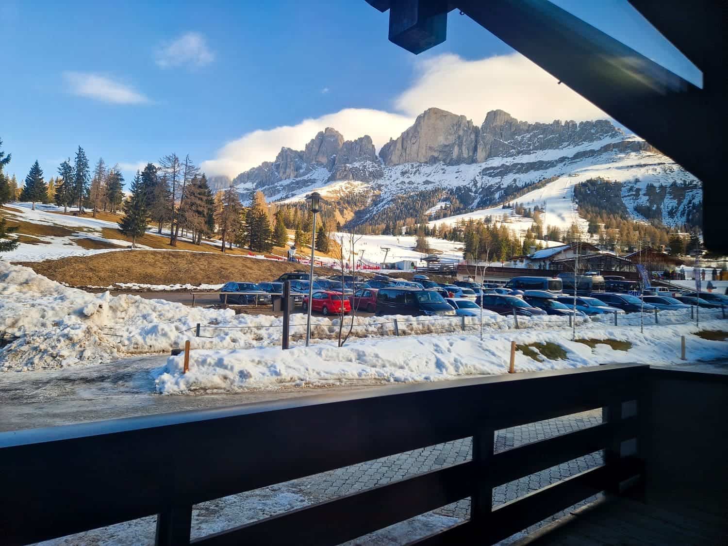 A scenic view of a snowy mountain landscape with rocky peaks in the background. Cars are parked on a snow-covered lot, bordered by trees. The foreground features a wooden balcony, with patches of snow scattered on the ground.