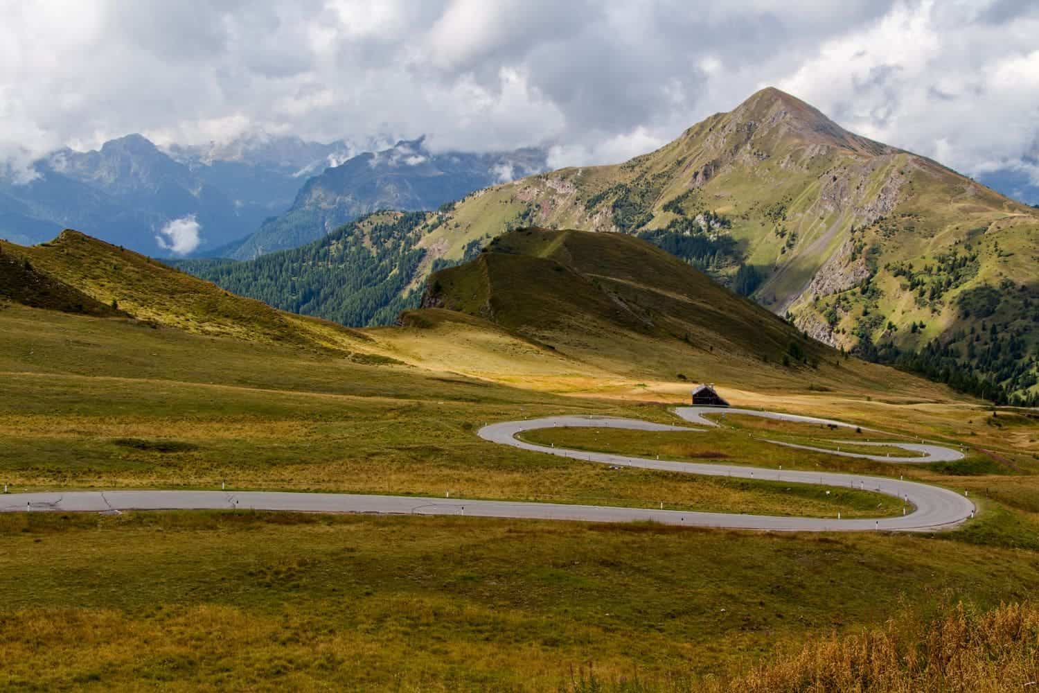 11 Short and Easy Hikes in the Dolomites: Unique and Must-do 23 A winding road curves through a lush green valley with mountains in the background under a cloudy sky. A small building stands near the road surrounded by grassy hills.