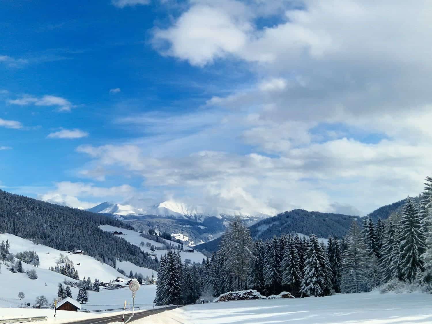 Are the Dolomites worth visiting? Yes! Why? 10 A scenic winter landscape showing snow-covered hills and pine trees under a blue, partly cloudy sky. Majestic mountains are visible in the distance, and a small wooden cabin sits near the foreground.