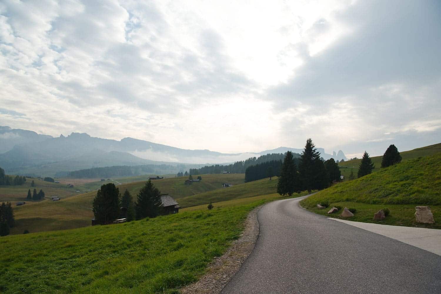 11 Short and Easy Hikes in the Dolomites: Unique and Must-do 12 A winding road curves through a lush green landscape with scattered trees and small cabins. In the distance, misty mountains rise under a partly cloudy sky, creating a serene and picturesque scene.