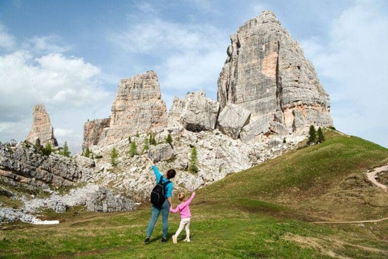 A woman and a child with raised arms stand on a grassy hill, facing towering gray rock formations under a partly cloudy sky. The scene suggests a feeling of exploration and awe in a natural mountainous landscape.