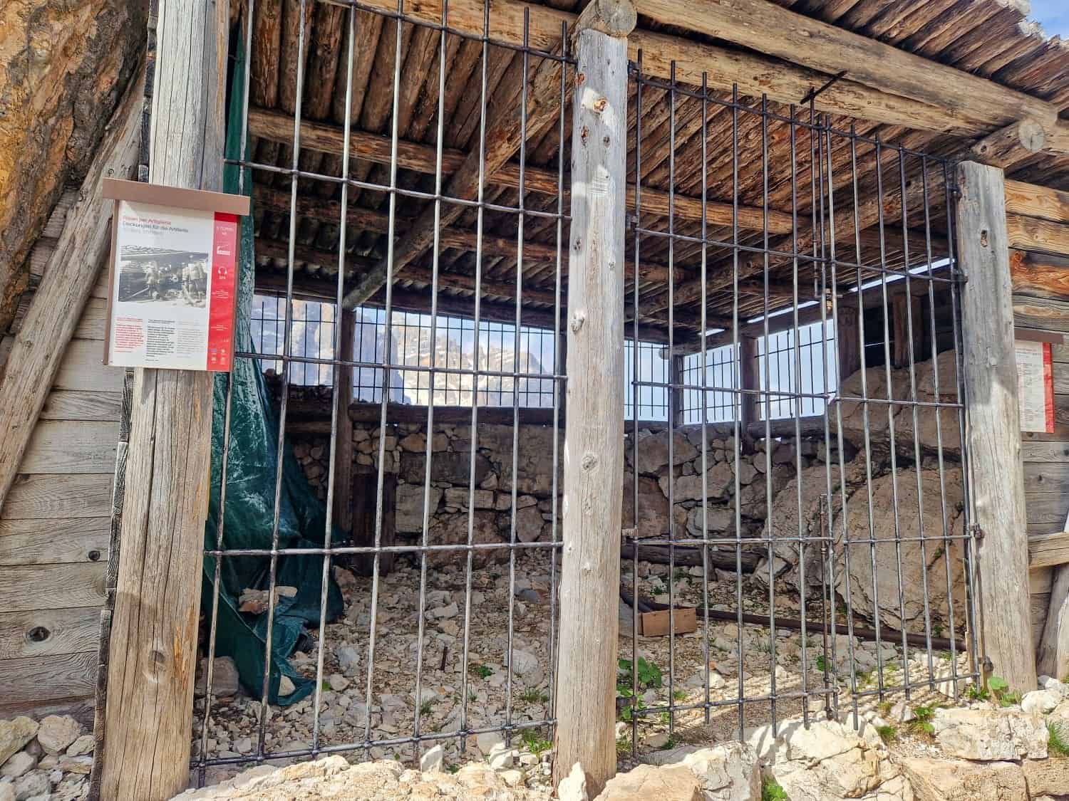 11 Short and Easy Hikes in the Dolomites: Unique and Must-do 14 A rustic wooden and stone shelter with a metal gate stands on a rocky terrain. Informational signs are placed on the wooden beams. The shelter's roof is made of overlapping wooden planks, and the interior appears empty.