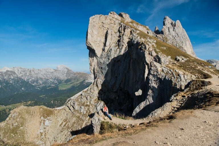 A person sits on a rocky path near a large jagged rock formation against a backdrop of distant mountains and a clear blue sky. The landscape features sparse vegetation and rugged terrain.