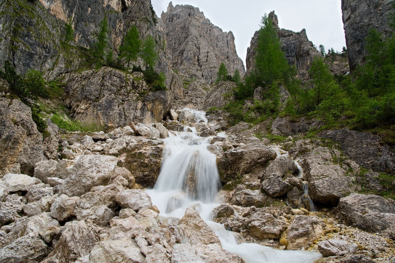 A stunning waterfall flows down a rocky mountainside surrounded by lush green trees and towering peaks. The water cascades over large boulders, creating a picturesque and serene natural scene.