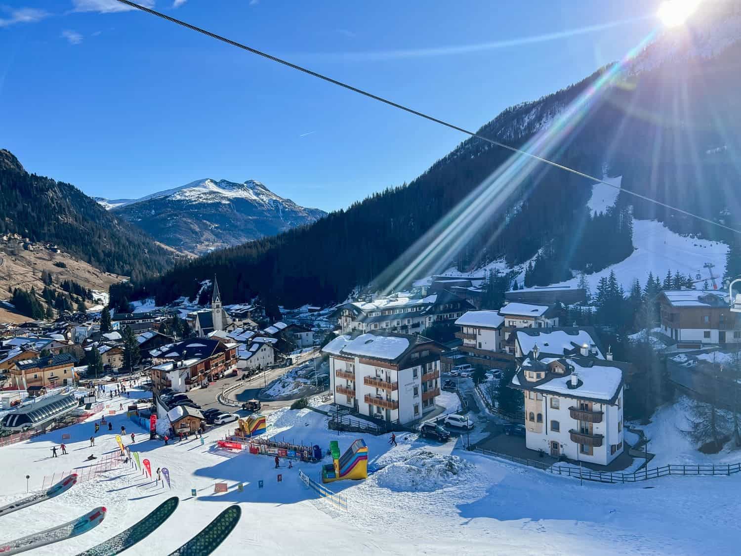 A picturesque ski resort village nestled in a snowy valley with a clear blue sky. Sunlight beams across the sky, casting light on the snow-covered chalets and a ski slope. Mountains rise in the background.