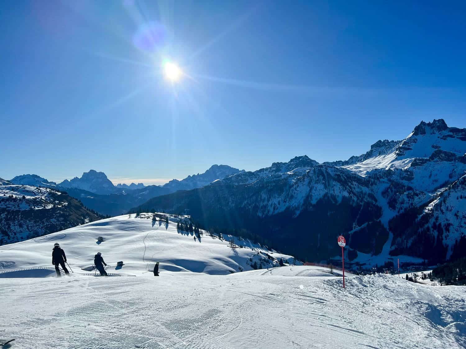 Skiers and snowboarders glide down a sunlit snowy mountain slope. The sky is clear and blue, with a bright sun shining above. Jagged, snow-covered mountain peaks rise in the background. A trail marker is visible.