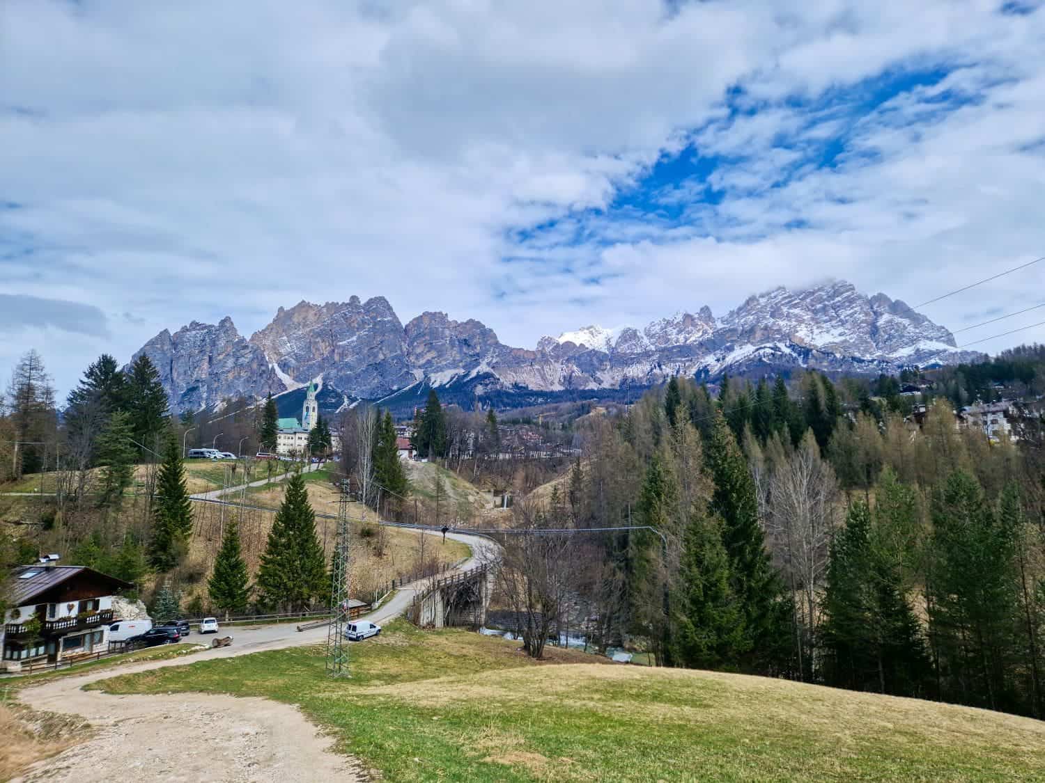 A scenic landscape features a winding road leading through a hilly area with sparse trees. In the background, rugged snow-capped mountains rise under a partly cloudy sky. A few houses and a small church are nestled among the trees.