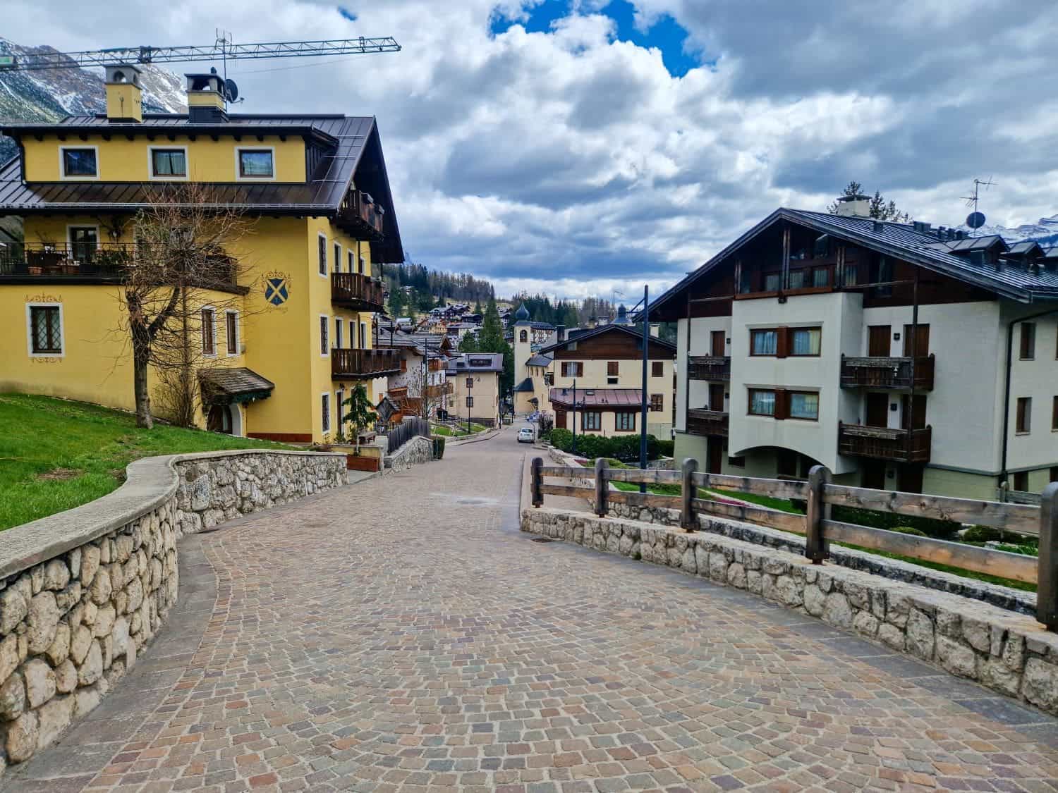 A cobblestone street in a quaint alpine village is lined with charming buildings featuring wooden balconies and pastel facades. Snow-capped mountains and a partly cloudy sky are visible in the background.