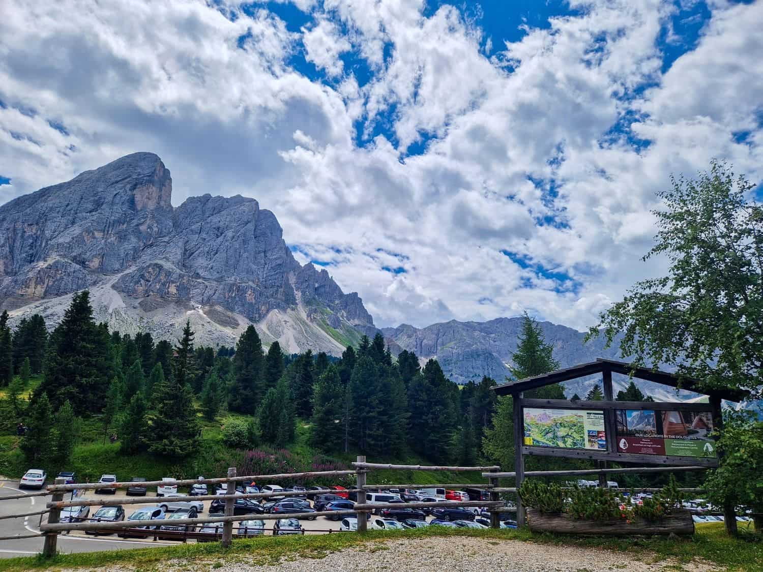 Scenic mountain view with rocky peaks and scattered clouds above. A parking area with numerous cars is surrounded by lush green trees. A wooden information board is in the foreground on the right.