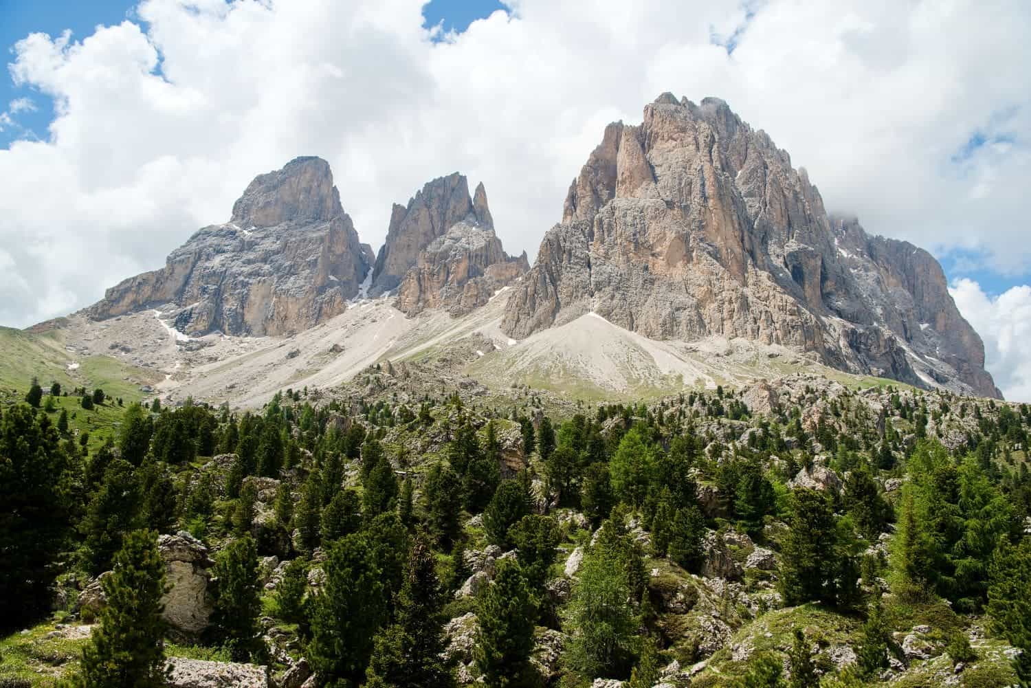 Majestic mountain peaks rise under a partly cloudy sky, with rocky slopes and a lush green forest in the foreground.