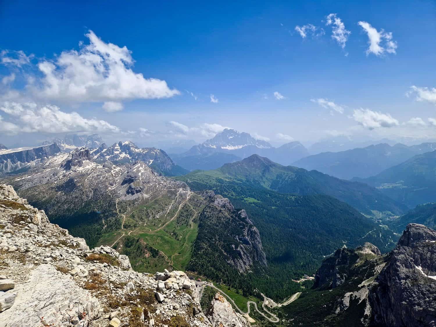 A breathtaking alpine landscape with jagged rocky peaks and lush green valleys under a clear blue sky. Snow-capped mountains are visible in the distance, with winding trails and scattered clouds enhancing the scenic view.