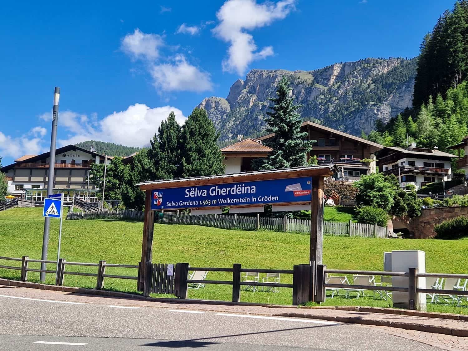 Mountain village scene with a sign reading "Selva Gherdëina" in front of traditional Alpine-style buildings. Lush greenery surrounds the area, with a clear blue sky and rocky mountain peak in the background.