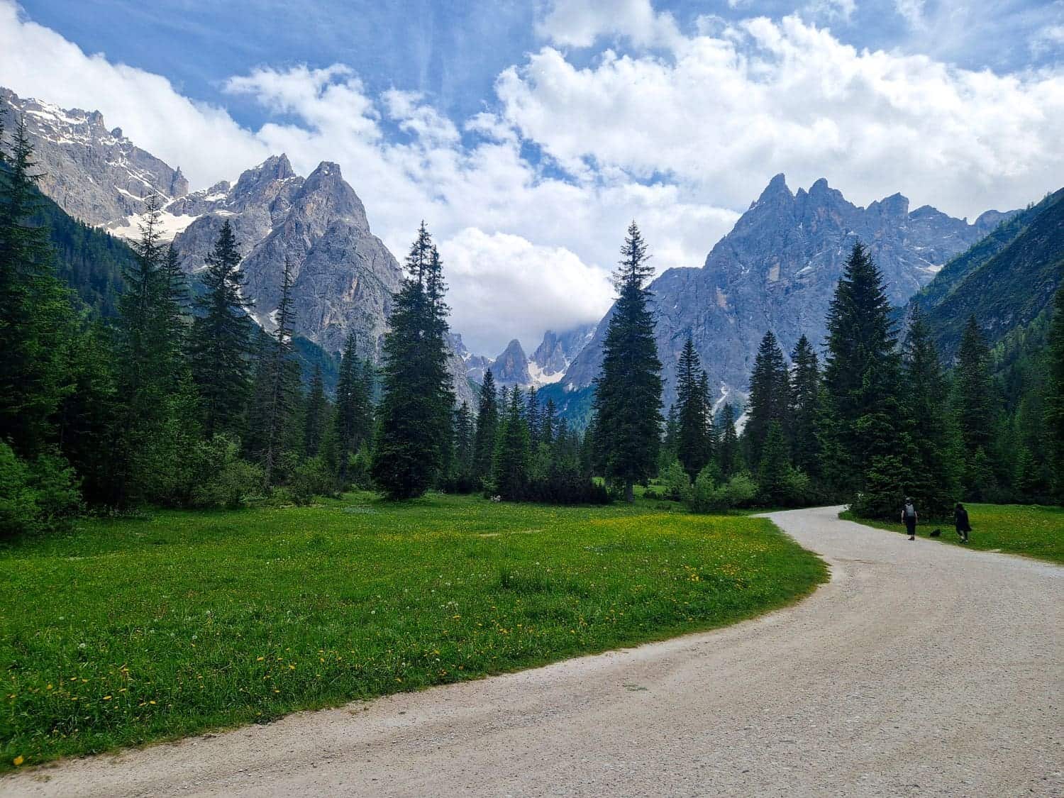 A scenic mountain landscape with towering peaks and patches of snow. A winding dirt path leads through a lush green meadow dotted with trees. Two people walk along the path under a partly cloudy blue sky.