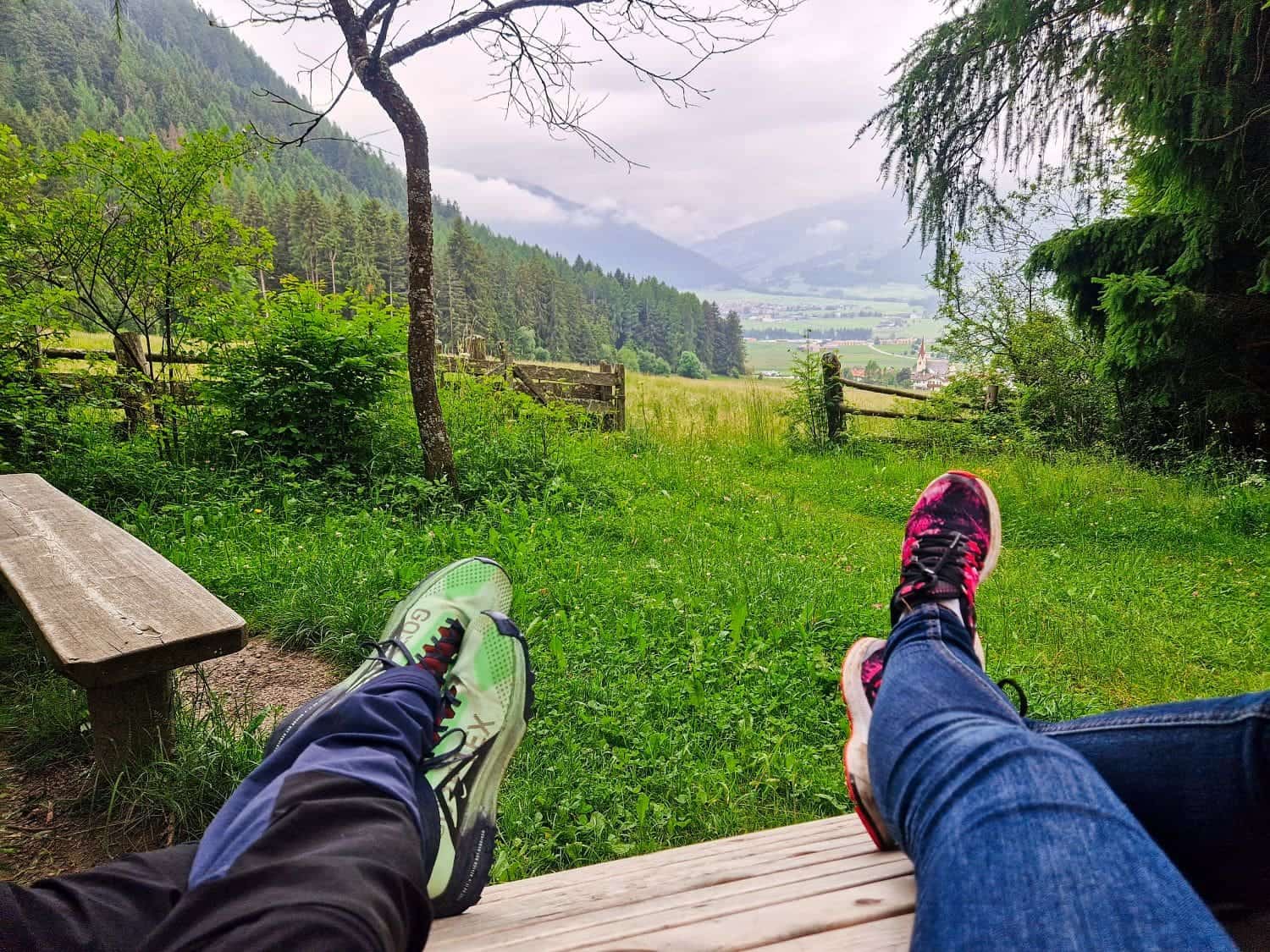 Two people relax on a wooden bench, overlooking a scenic valley with green fields and distant mountains. They wear athletic shoes—one green and black, the other pink and black—and jeans, surrounded by lush trees and a cloudy sky.