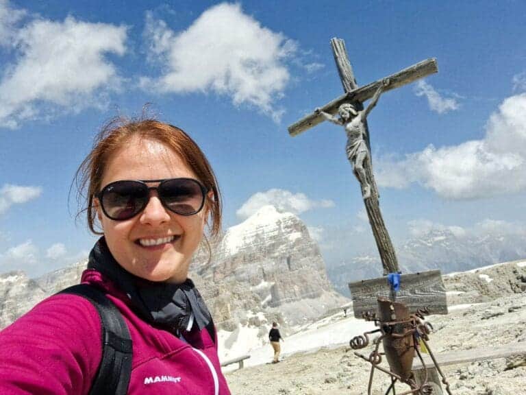 A woman wearing sunglasses and a maroon jacket smiles for a selfie on a snowy mountain peak. Beside her is a wooden crucifix with a statue of Jesus. The background shows a partly cloudy sky and distant mountains.