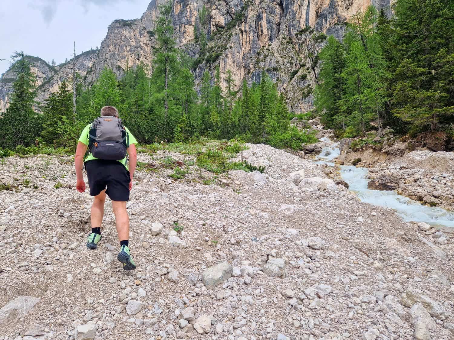 A person wearing a backpack hikes on rocky terrain near a flowing creek. Tall trees and steep, rugged cliffs are visible in the background under a cloudy sky.