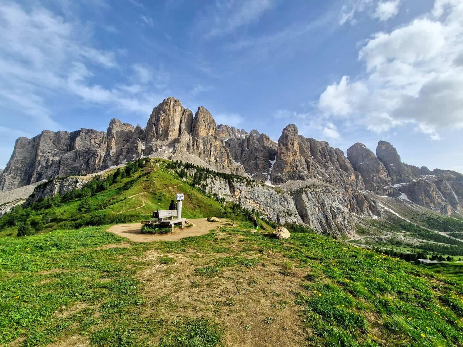 A scenic view of the Dolomites with rugged mountain peaks under a partly cloudy sky. A small building sits on a green hill with a winding path surrounded by lush grass and vegetation in the foreground.
